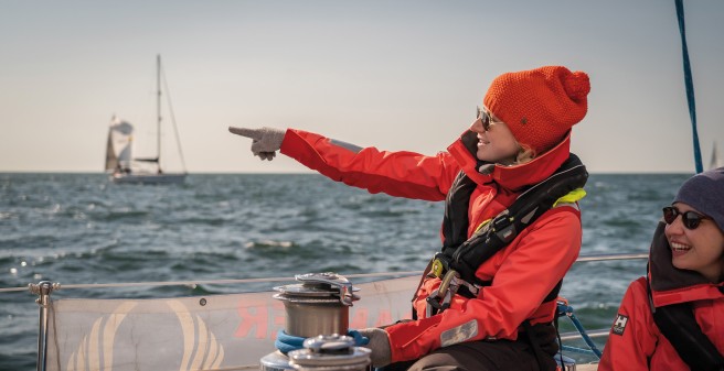 Segelrebellen, eine rotgekleidete blonde Frau zeigt mit gehobenem Arm auf etwas in der Ferne