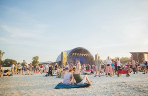 Blauer Himmel, Blick auf das Festival Gelände, die Bühne klein im Hintergrund, Menschen unterschiedlichen Alters sitzen und stehen lässig beieinander