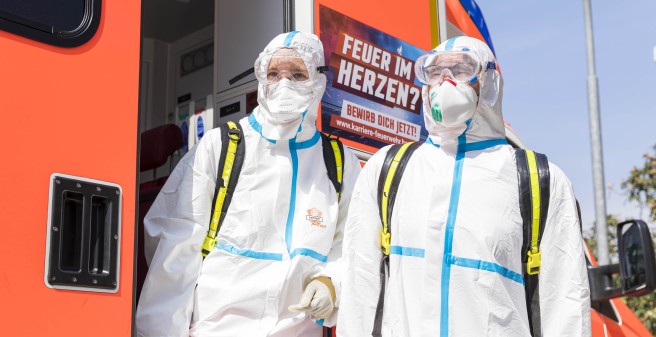 Notäztin Stefanie Beck  und ihr Kollege, Notfallsanitäter Sebastian Götsch, steigen aus dem rotorange farbenen Krankenwagen aus. Sie sind in den Schutzanzügen und mit den Masken komplett verhüllt. Der Himmel ist strahlend blau.