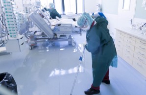 Blick in ein Zimmer der Intensivstation. Sehr weiß, sehr hell. Im Hintergrund ein einzelner Patient im Bett liegend, das Oberteil des Bettes ist hoch gestellt. Im Hintergrund eine Pflegekraft. Vorn wischt eine Frau in Schutzkleidung den länzenden Boden.