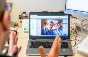 Blick über die Schulter von Herrn Prof. Buhmann. Vor ihm ein aufgeklappter Laptop, auf dem Bildschirm des Gerätes sind zwei ältere Personen zu sehen. Der Mann und die Frau blicken Kopf an Kopf in die Kamera.