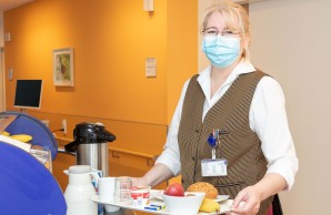 Bianca Stuchlik, mit Mund-Nasen-Schutzmaske lächelt in die Kamera. Sie hat ein Frühstückstablett mit einer appetitlichen Mischung aus Brötchen, Belag und Obst zusammengestellt.