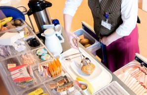 EIn Blick auf das Frühstücksbüffet der Patienten. Eine Mitarbeiterin stellt einen Teller mit Brötchen und frischem Obst zusammen