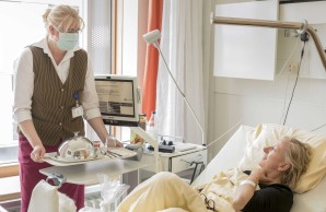 Bianca Stuchlik, eine blonde Frau, die Haare zum Zopf gebunden, mit Mund-Nasen-Schutz, steht vor dem Bett einer Patientin. Diese liegt im Bett und lächelt die UKE Mitarbeiterin an. Frau Habermann stellt ein Tablett auf den Beistelltisch des Krankenbetts. Das Gericht ist mit einer glänelnden Glosche abgedeckt.