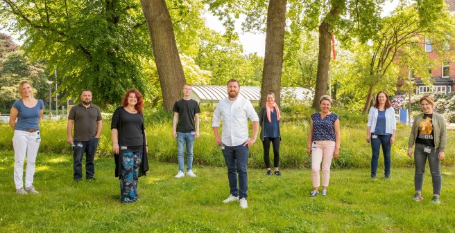 Neun Frauen und Männer stehen auf einer Wiese und grünen, höhen Baumen. Sie lächeln und stehen entspannt. Ein sonniger Tag. Die einzelnen Personen der Gruppe haben einen großen Abstand zueinander