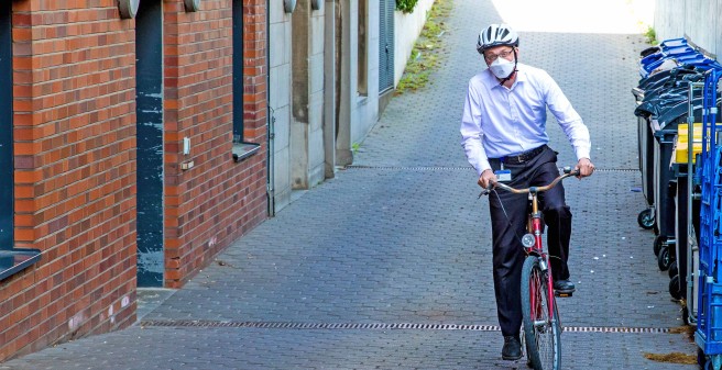 Sven Gerber, in dunkelblauer Tuchhose, hellblaubem Hemd, Fahrradhelm und Atemschutzmaske radelt auf einem roten, etwas rotigem Rad eine leichte Steigung. der Weg ist gepflaster, links GEbäudeeingänge, rechts Müllcontainer.Er blickt angestrengt.