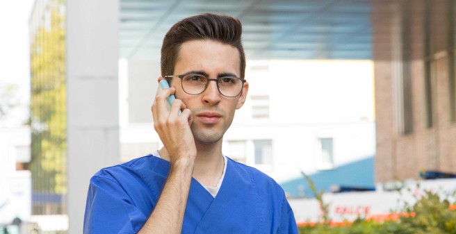 Christopher Sila, ein junger Mann mit dunklen, kurzen Haaren und dunkler Horbrille, blickt in die Kamera, er hält ein Handy ans Ort. Im Hintergrund die Notaufnahme