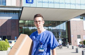 Christopher Silva in blauer ARbeitskleidung, steht vor dem Haupteingang des UKE, unter dem Arm trägt er ein großes Paket