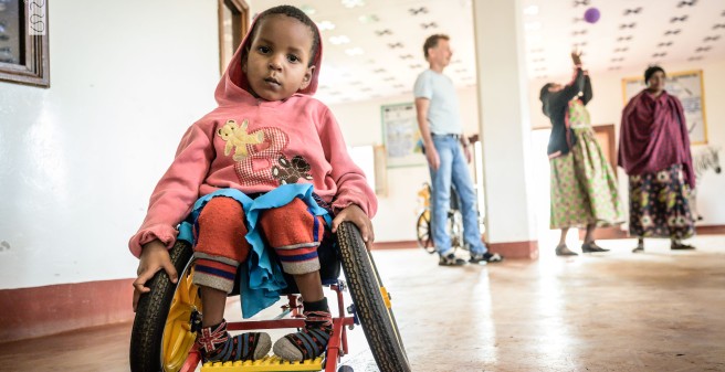 ein afrikanisches Kind im Rollstuhl im Klinikgebäude, im HIntergrund drei Erwachsene