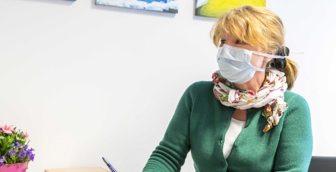 Beate Gerber, Lob- und Beschwerdemanagement im Gespräch mit Patientin Frau Trieb. Frau Gerber, mit Atemschutzmaske, trägt eine grüne Strickjacke und Halstuch mit Blumenmustern. Sie sitzt am Tisch, ein Kugelschreiber in der Hand und blickt ihr Gegenüber an.