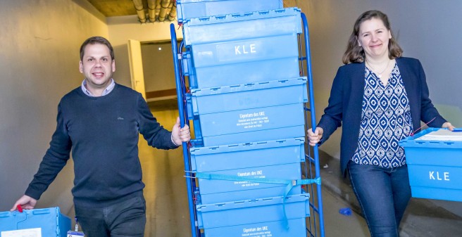 David Bielang, Laura Weinhold schieben einen Rollwagen durch einen Liefergang des UKE, der Wagen ist voll beladen mit blauen Kisten