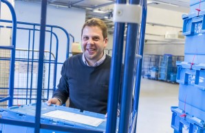 Daniel Bielang, in gemustertem Hemd und blauem Pullover, schaut lächelnd in die Kamera. Er stellt gerade eine blaue Kiste in einen Rollconteiner. Rechts von ihm ein hoher Stapel weiterer Kisten. 