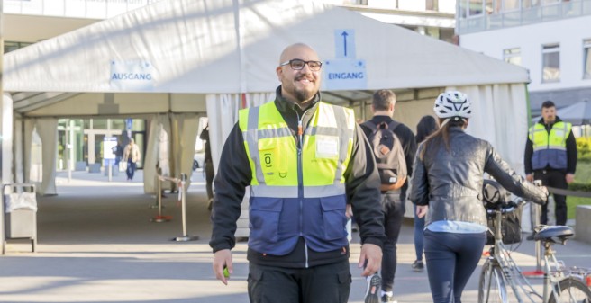 Ein Mitarbeiter des Sicherheitsdienstest, in gelber Warnweste, steht vor einem Zelt für den Ein- und Ausgang. Eine Mitarbeiterin mit Fahrrad darf passieren.