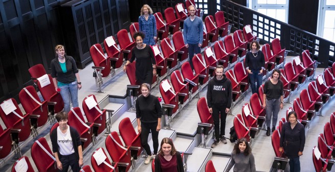 Blick von oben auf ein vielkopfiges Team aus  UKE und Bernhard-Nocht-Institut für Tropenmedizin Mitarbeitern, darunter Prof. Dr. Marylyn  Addo. Sie stehen im Hörsaal mit großem Abstand zueinander,, die Sessel sind gem. Coronabestimmungen mit weißen Bändern gesperrt, Schilder mit Namen zeigen die Platzverteilung.